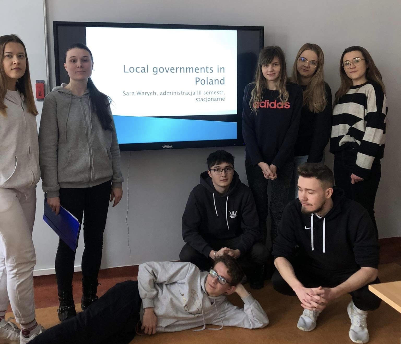 Grupa ośmiu młodych ludzi stoi, klęczy lub leży przed tablicą interaktywną. Na tablicy wyświetlony jest napis: Local governments in Poland, Sara Warych, administracja III semestr, stacjonarne.